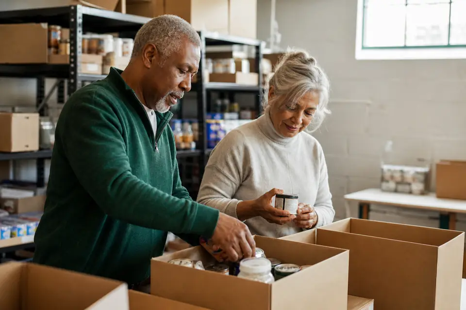 Two volunteers packing supplies into boxes in a half-empty food pantry storeroom.